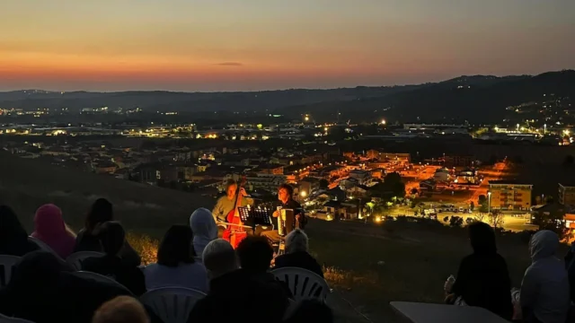 Musicisti con violoncello e fisarmonica in concerto notturno a Montelabbate, pubblico e borgo illuminato sullo sfondo